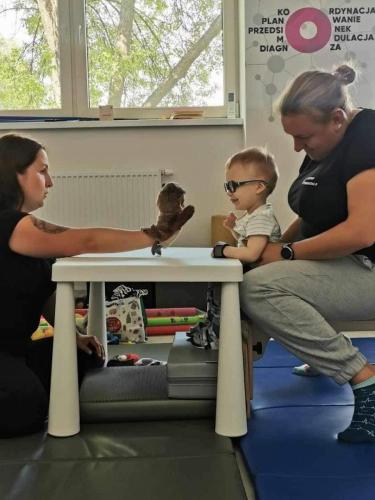 Mały chłopiec w okularach obserwuje pacynkę prezentowaną przez terapeutkę podczas ćwiczeń rozwijających koncentrację wzrokową i uwagę. 