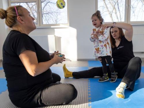 Fizjoterapeutki wspierają dziewczynkę podczas rehabilitacji funkcjonalnej, ćwicząc równowagę, stabilizację i pozycję stojącą. 