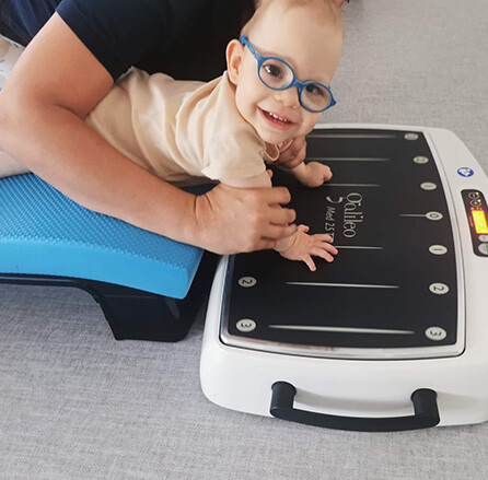 Uśmiechnięte dziecko w okularach podczas terapii ruchowej na platformie wibracyjnej Galileo, wspierane przez terapeutę w trakcie zajęć rehabilitacyjnych w Centrum Grawitacja.